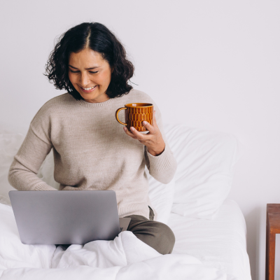 Woman sitting on her bed looking at her laptop holding a coffee cup